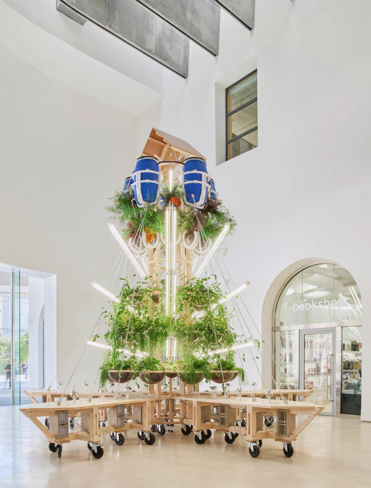 CON-VIVERE, The Edible Tower, is an architectural display that stands covered in natural materials like draping leaves and wooden frames in a light-filled gallery.